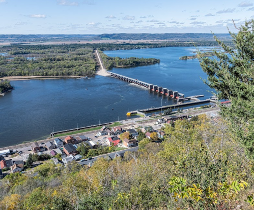 Aerial view of a river with a dam, bridge, and a small town along the shore, surrounded by trees and hills.