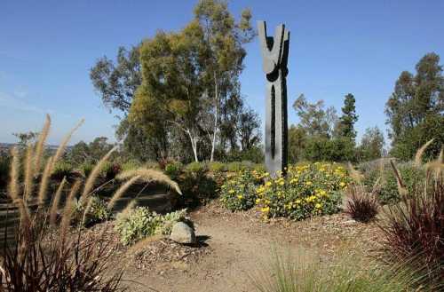 A tall, abstract sculpture surrounded by colorful flowers and greenery under a clear blue sky.