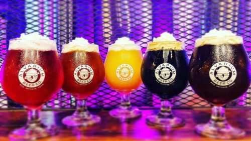 Five glasses of beer in various colors, topped with foam, displayed against a colorful, illuminated background.