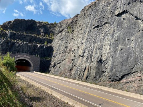 A road leading into a tunnel carved into a rocky cliff, surrounded by steep, rugged terrain and blue sky.