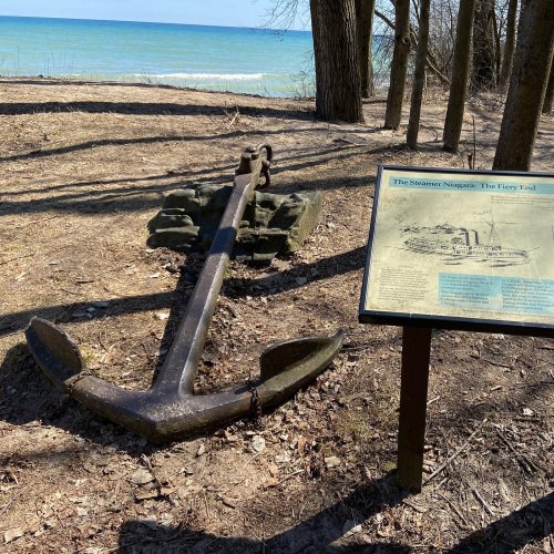 An old anchor rests on the ground near a lake, with a sign about the steamer Niagara in the background.