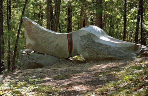 A large, abstract stone sculpture resembling a whale, nestled among trees in a forested area.