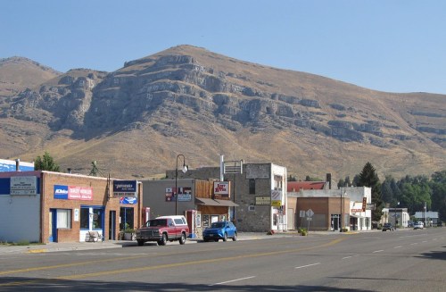 A small town street with shops and vehicles, set against a backdrop of dry, mountainous terrain.