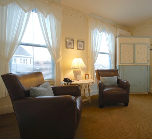Cozy living room with two brown armchairs, a small table, a lamp, and large windows with white curtains.