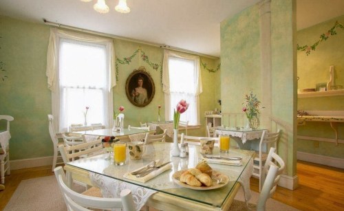 Cozy dining room with green walls, tables set for breakfast, flowers, and a portrait on the wall.