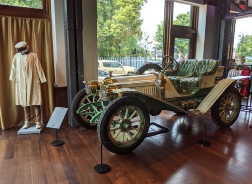 A vintage car displayed indoors next to a mannequin in traditional clothing, surrounded by large windows and wooden flooring.