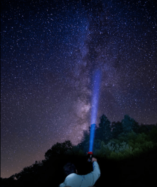 A person points a flashlight at the starry night sky, revealing the Milky Way above a forested landscape.