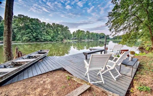 A serene lakeside view featuring a wooden dock, chairs, and a small boat, surrounded by lush greenery and calm water.