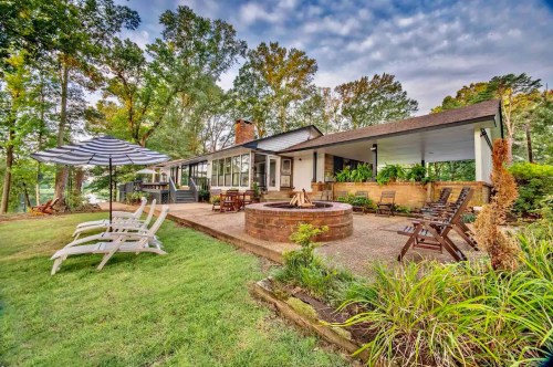 A cozy lakeside home with a fire pit, patio chairs, and an umbrella, surrounded by lush greenery and trees.