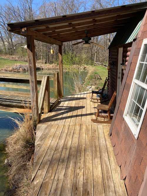 A wooden deck beside a pond, featuring a rocking chair and a covered area with a fan, surrounded by trees.