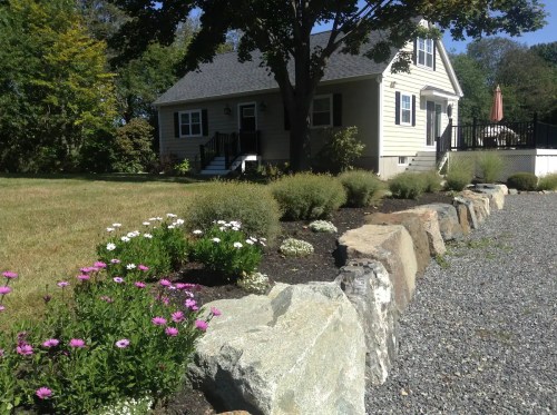 A cozy house surrounded by a landscaped yard with flowers and a stone border, under a clear blue sky.