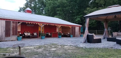 A rustic barn with a red exterior, surrounded by greenery, features outdoor seating and a gazebo on a gravel path.