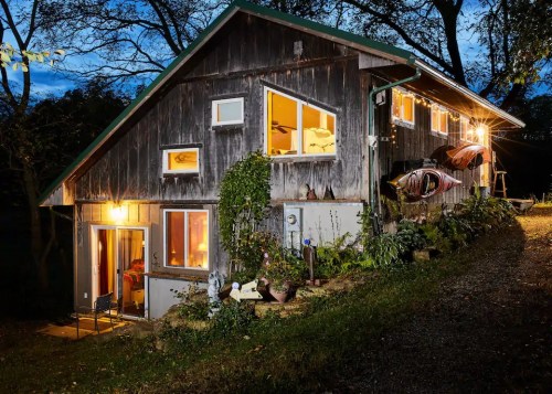 A rustic wooden house at dusk, with warm lights glowing from the windows and kayaks displayed on the exterior.