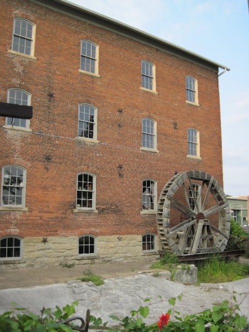 Historic brick building with large wooden water wheel and multiple windows, surrounded by greenery.