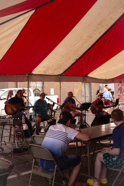 A band performs under a red and white striped tent, with an audience seated at tables in the foreground.