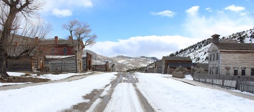 A snowy, deserted street in a ghost town, flanked by old wooden buildings and mountains under a blue sky.