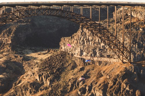 Two wingsuit flyers glide near a large bridge over a rocky canyon landscape during sunset.