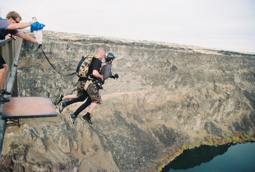 Two people jump off a platform into a canyon, with one person holding onto the other, surrounded by spectators.
