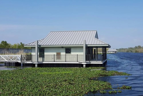 A modern building on stilts surrounded by water and greenery, under a clear blue sky.