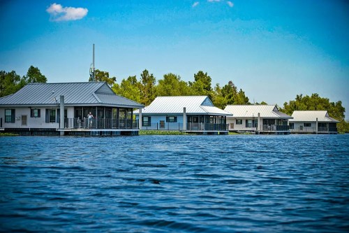 A row of waterfront houses with metal roofs, surrounded by trees and reflecting in the calm water under a blue sky.