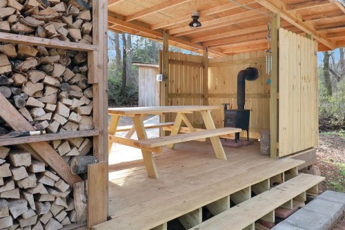 A wooden outdoor shelter with a picnic table, woodpile, and a small stove, surrounded by trees.