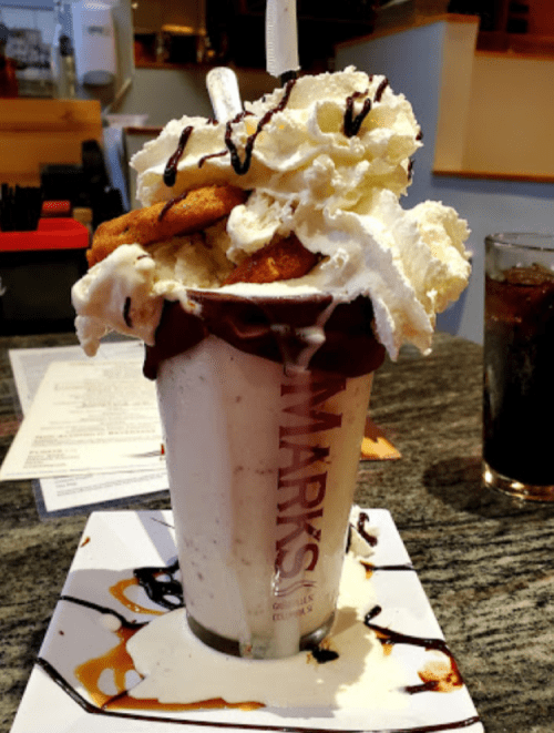 A tall milkshake topped with whipped cream, cookies, and chocolate drizzle, served on a plate with caramel sauce.