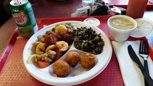 A plate of fried chicken, shrimp, collard greens, and cornbread, with a can of ginger ale on a red tray.