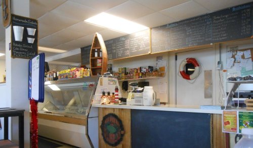 Interior of a small deli with a counter, menu boards, and shelves stocked with various food items.