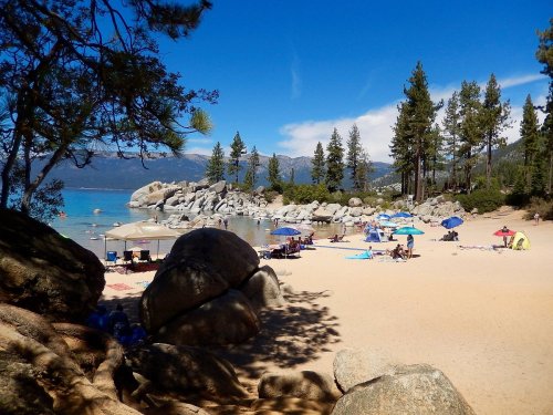 A sandy beach by a lake, surrounded by trees and rocky formations, with people relaxing under umbrellas on a sunny day.