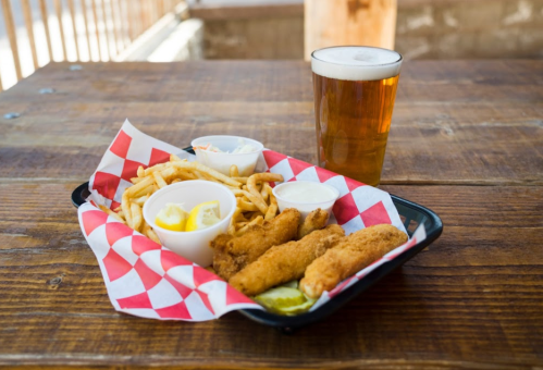 A tray with fried fish, fries, coleslaw, and dipping sauces, alongside a glass of beer on a wooden table.