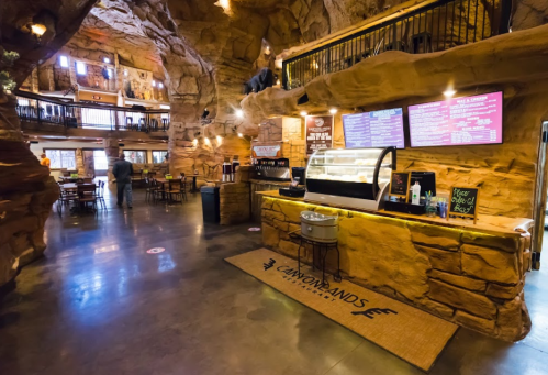 Interior of a rustic café with rock formations, wooden tables, and a counter displaying food and drinks.