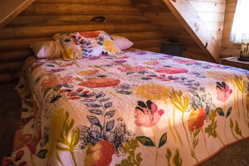 A cozy attic bedroom with a floral quilt on a bed, surrounded by wooden walls and soft lighting.