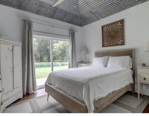 Bright bedroom with a wooden bed, white linens, and large windows overlooking a green outdoor space.