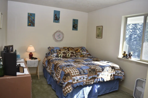 Cozy bedroom with a blue quilted bedspread, wall art, a lamp, and a window letting in natural light.