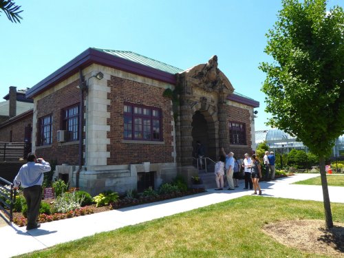 A historic brick building with a decorative entrance, surrounded by greenery and people enjoying the outdoor space.