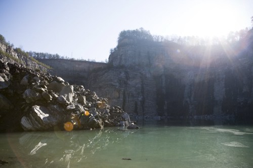 A serene landscape featuring a calm, greenish lake surrounded by rocky cliffs and sunlight filtering through trees.