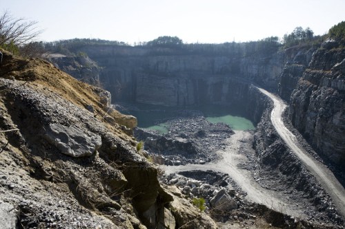 A deep quarry with steep rocky walls and a small body of green water at the bottom, surrounded by a dirt road.