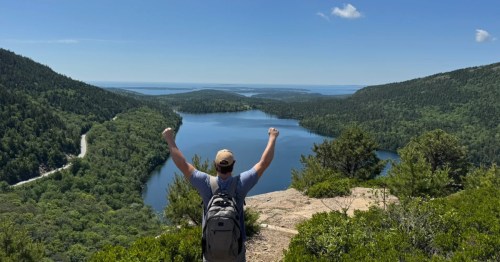 A man extends his hands in a victory pose as he admires a spring hike in Maine.