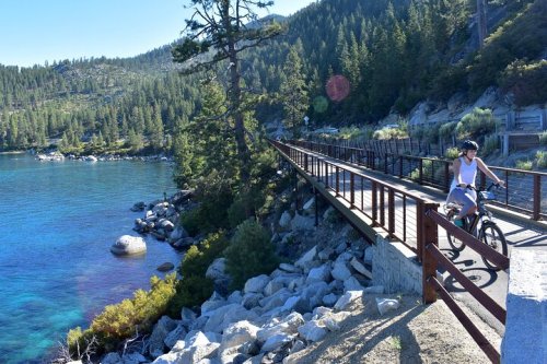 A cyclist rides along a scenic path by a clear blue lake, surrounded by trees and rocky shores.