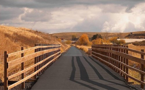 A scenic pathway lined with wooden fences, surrounded by golden fields and a cloudy sky.