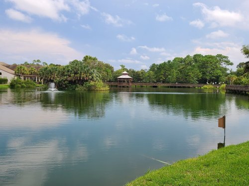 A serene lake surrounded by greenery, featuring a gazebo and a small waterfall under a blue sky with fluffy clouds.