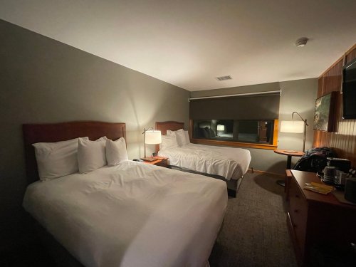 A dimly lit hotel room featuring two beds, a window, and a small desk with a lamp and coffee maker.