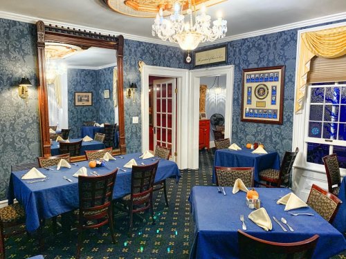 Elegant dining room with blue tablecloths, neatly set tables, and ornate decor, featuring a chandelier and large mirror.