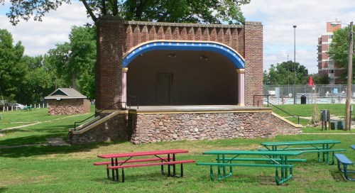 A brick outdoor stage with colorful accents, surrounded by green grass and picnic tables in a park setting.