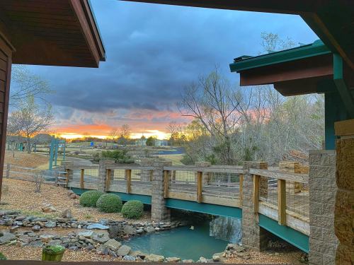 A wooden bridge crosses a small stream, framed by trees and a colorful sunset under a cloudy sky.