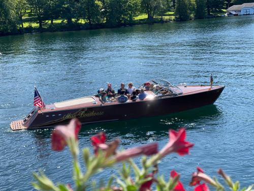 A classic wooden boat with passengers cruising on a serene lake, surrounded by greenery and flowers in the foreground.