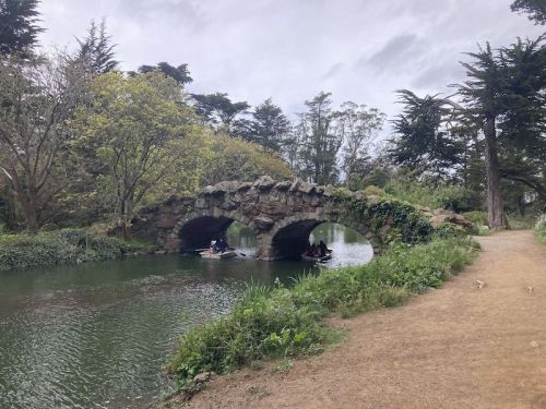 A stone bridge arches over a calm river, with kayakers paddling beneath and lush greenery lining the banks.