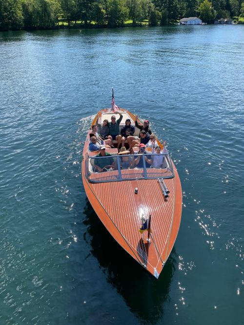 A group of people enjoying a sunny day on a wooden boat in a calm lake surrounded by greenery.