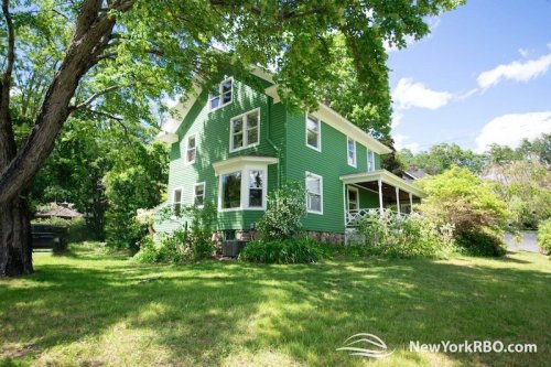 A green two-story house surrounded by trees and a grassy yard under a clear blue sky.