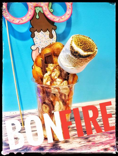 A colorful dessert cup filled with popcorn, donuts, chocolate, and a toasted marshmallow, with a playful "BONFIRE" text.